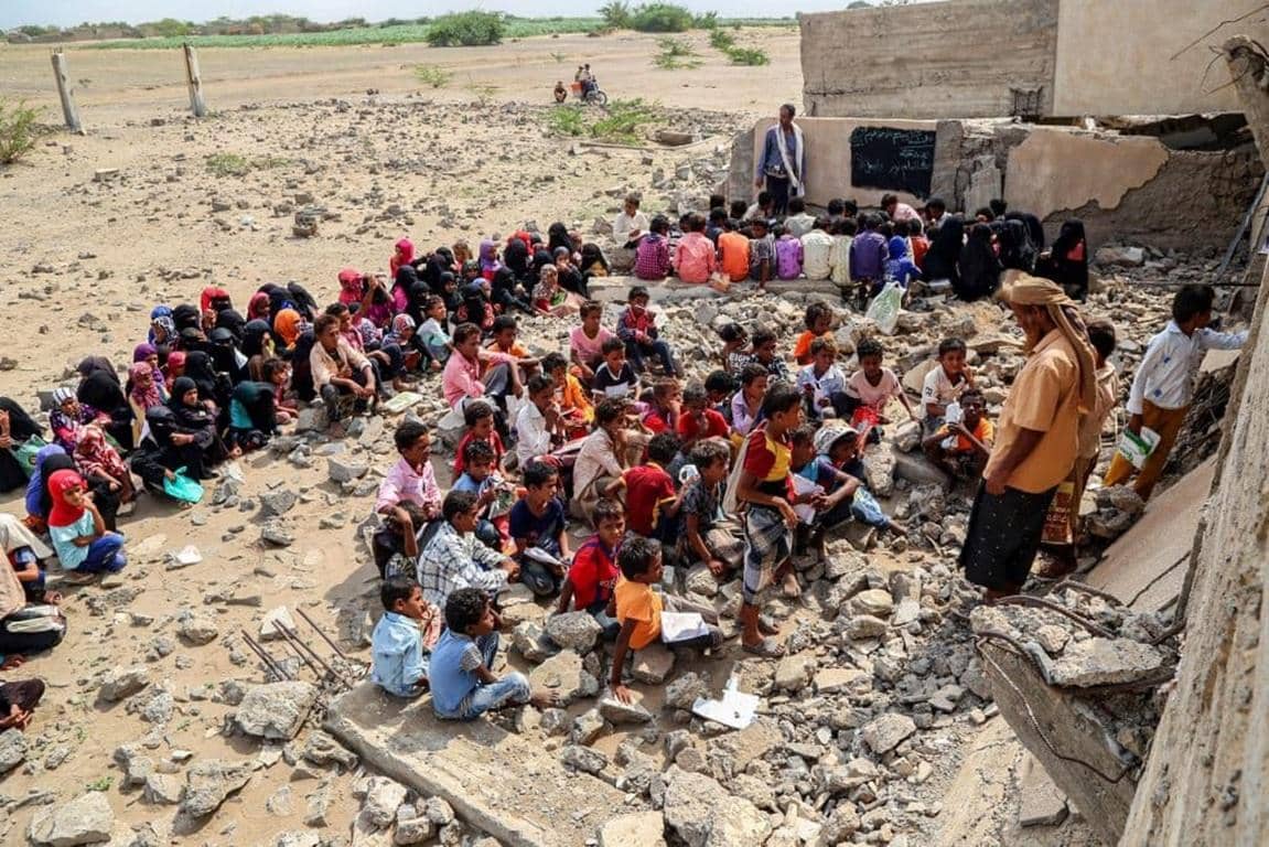 Children sit on rubble in front of a damaged structure, a teacher stands to address them, representing hope and education in areas supported by crypto donations like ETH.