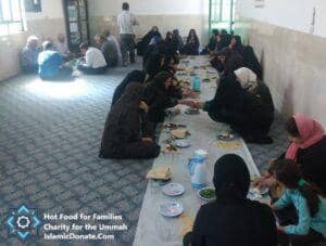 Group of people, some men and women, sitting on the floor gathered around a long table for a meal. This event is supported by crypto donations, providing hot meals for families. Key elements: daily charity, relief for the needy, sustainable livelihoods, hot meals, Islamic relief, aid provision, with support via ETH.