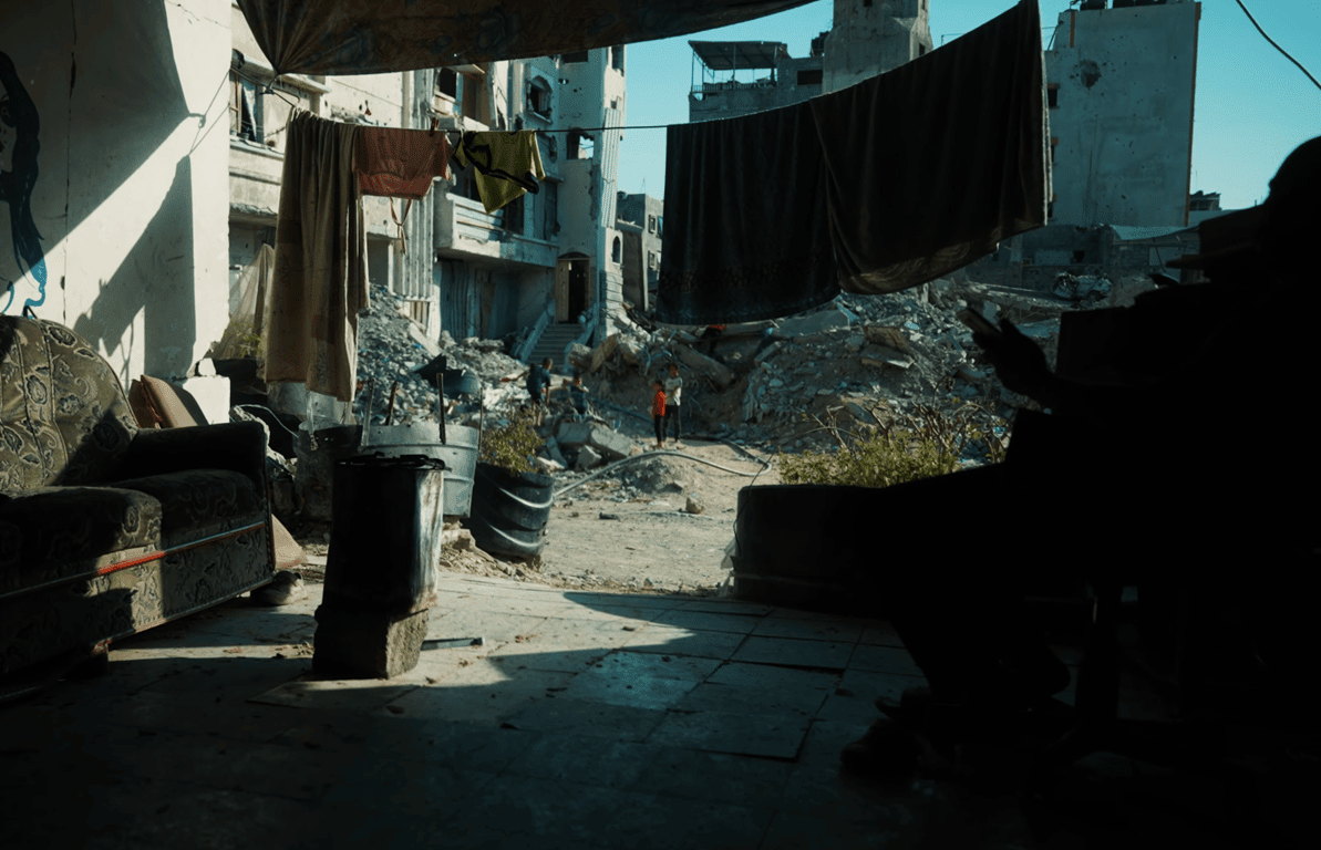 Children stand amidst rubble and destroyed buildings in Gaza; a ruined home with a sofa and clothesline suggests ongoing displacement, with aid facilitated by BTC donations.