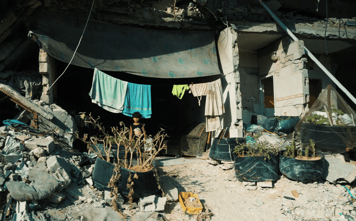 Child stands near laundry hung out to dry amidst rubble. Refugee camp, Gaza. Donate via ETH for urgent aid.