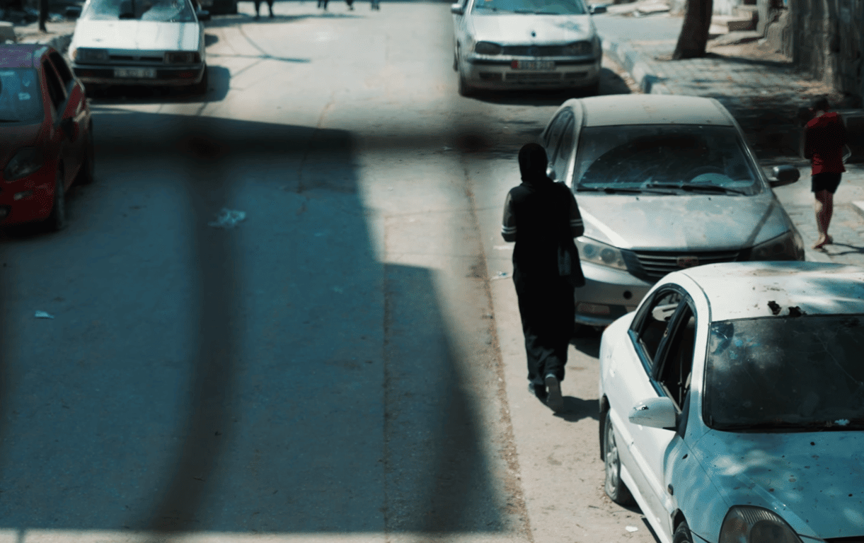 A woman in a headscarf walks down a street lined with parked cars. This street scene from Gaza reflects the urgent needs of refugees, with donations in BTC facilitating critical aid like food, water, and shelter.