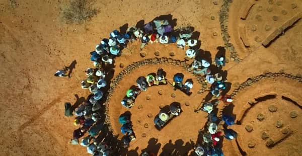 Aerial view of a diverse group of people gathered in a circular formation, participating in a tree planting or reforestation project in a dry, arid landscape. Donations support environmental protection initiatives, including reforestation efforts, with a nod to crypto donations like SOL.