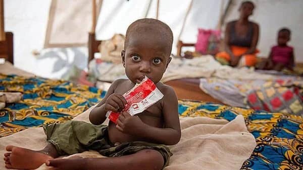 A child eats from a food packet in a tent, with adults blurred in the background. Help provide essential aid in Eritrea, like food, water, and vaccines, through cryptocurrency donations, supporting vulnerable populations with the help of Bitcoin.