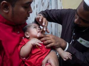 Tahnik ritual with baby receiving a sweet substance rubbed on the palate by a man in a brown thobe, while another man in a red outfit holds the baby. Traditional Islamic welcome with dates and prayer, supported by crypto donations.