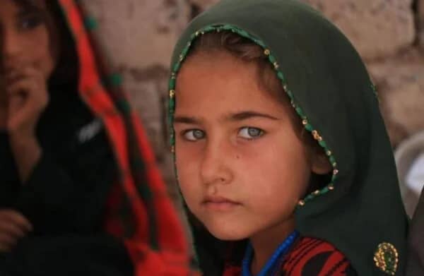 A young Afghan girl with striking blue eyes looks out with a serious expression. She wears a green hijab. This image represents children in need, vulnerable protection, and education support in Afghanistan.