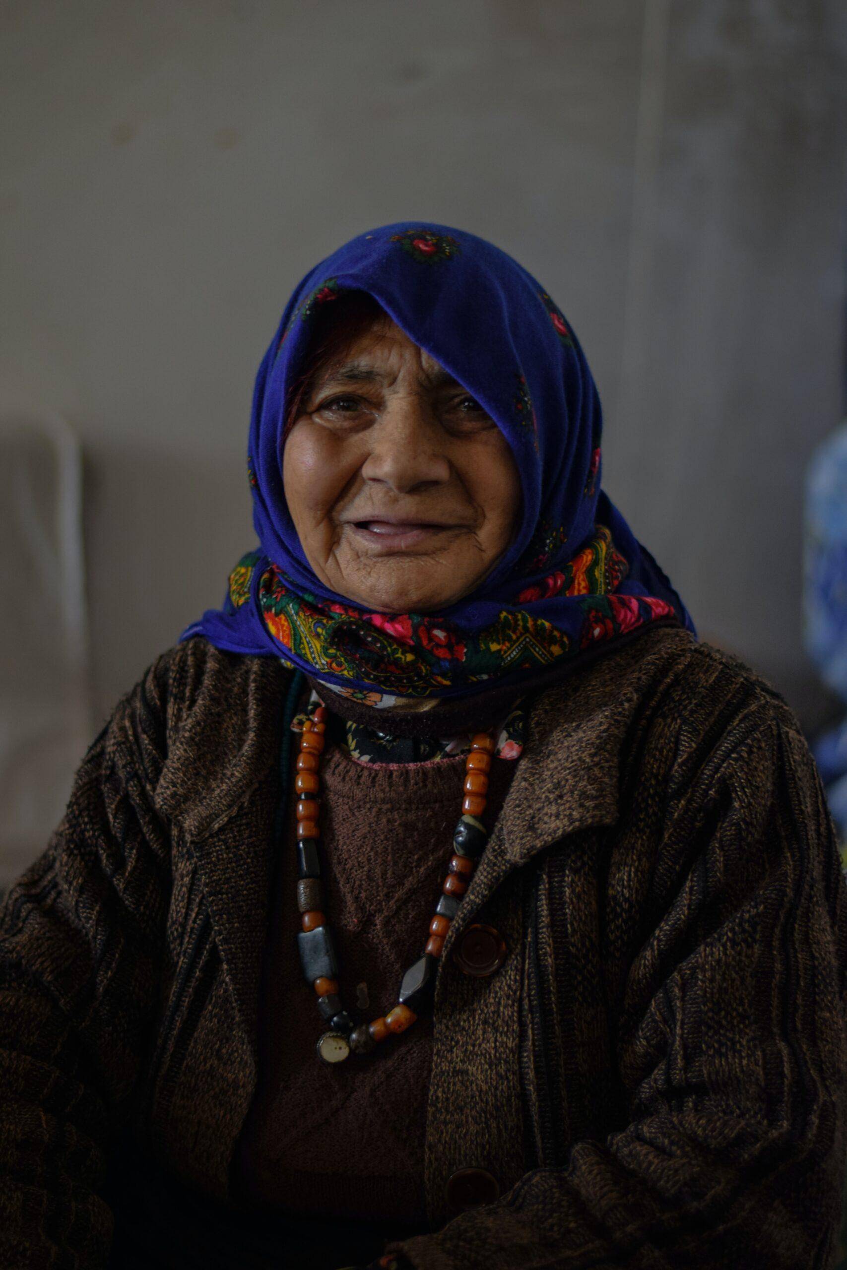 Elderly woman wearing a blue headscarf and traditional clothing, expressing hardship. Supported by crypto donations processed on BTC.