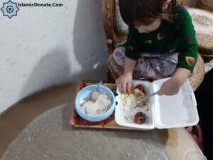 A child eats rice with tomatoes from a take-out container; rice is also in a blue dish. This act of giving is facilitated by IslamicDonate.com and supports crypto donations like BTC, providing nourishment for those in need as part of Aqiqah Qurbani for newborn babies.