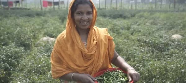 Aisha, an empowered woman in rural Bangladesh, holds a basket of fresh crops signifying the success of her farm, made possible by crypto donations and community support. Her story exemplifies how empowering women leads to flourishing families. Donation via XRP.
