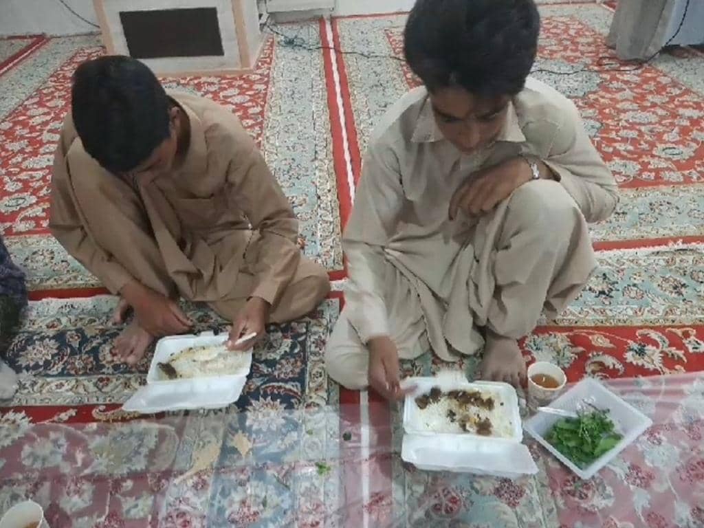 Two boys eating food from styrofoam containers on a patterned carpet. The scene depicts the distribution of meals, possibly for Kaffarah, with financial aid from crypto, such as BTC, supporting those in need.