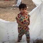 A young child stands amidst makeshift shelter fabric, symbolizing the ongoing daily struggles and resilience in Palestine. Aid delivery is hampered by checkpoints, waste management issues, and water scarcity. Supporting this cause with BTC donations helps overcome obstacles in delivering essential resources and education.