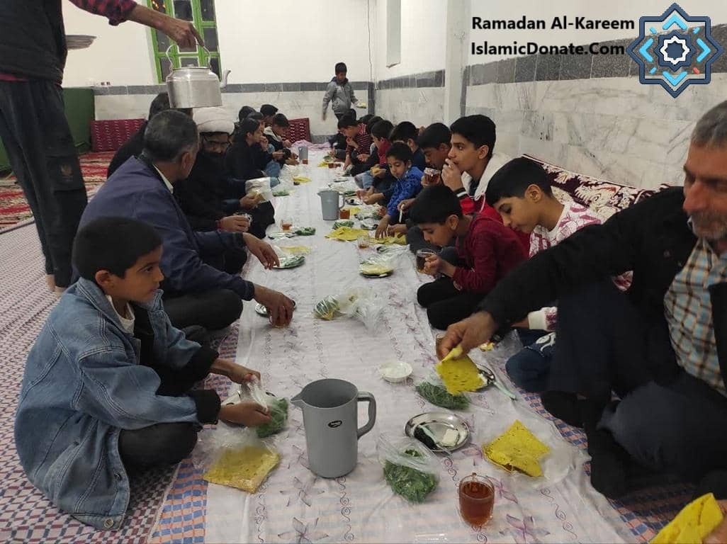 Children and adults sharing an Iftar meal, seated on the floor with food laid out on a large cloth. A person pours from a kettle, serving drinks. Crypto donations fund these charitable meals for the Muslim community, with contributions supporting Iftar packages and food hall distributions for those in need, particularly in conflict zones. The generosity facilitates breaking the fast with nutritious food, offering sustenance and hope.