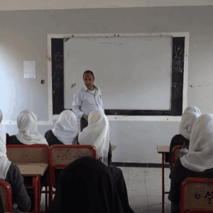 A teacher stands at a whiteboard in a classroom as students, mostly girls wearing headscarves, sit at desks. Sponsored education for 49 orphans, funded by crypto donations like BTC, provides hope and resources for brighter futures.