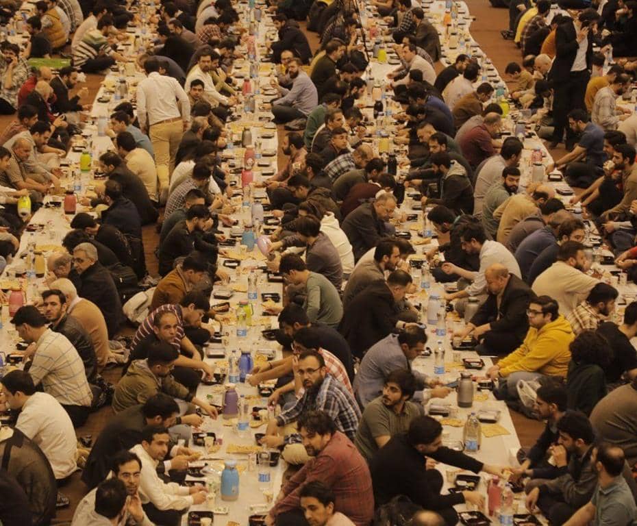 A large group of men sit cross-legged on the floor, breaking their fast together at long tables laden with food. Their communal Iftar, facilitated by crypto donations, spreads to war-torn regions like Palestine and Gaza, offering sustenance and respite during Ramadan.