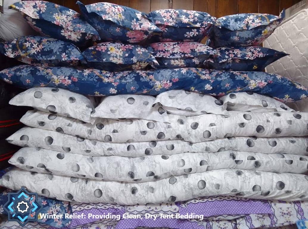 Stacked bedding including pillows and blankets featuring floral and polka dot patterns. These items provide essential winter relief for displaced families, made possible by crypto donations like BTC.