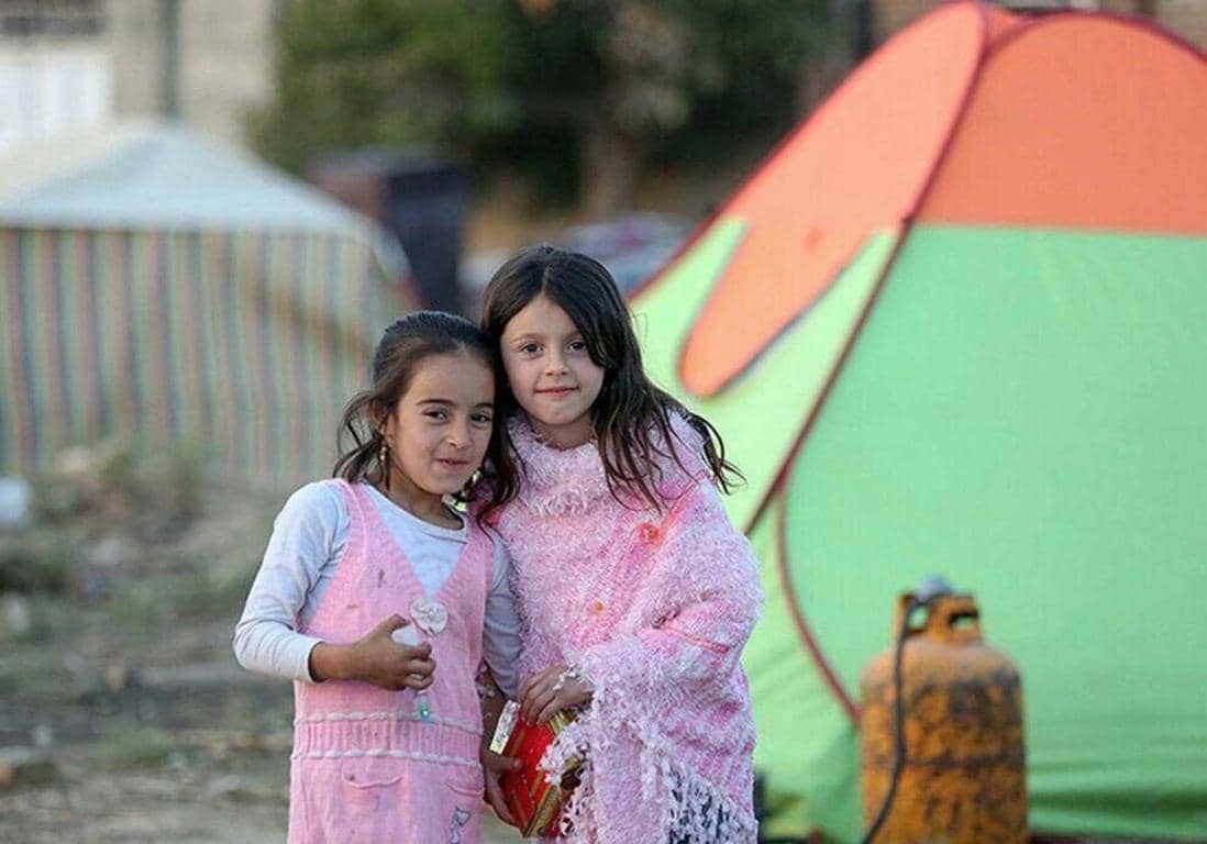 Two young girls in pink winter clothing stand in front of a tent, symbolizing the need for winter relief aid. Your crypto donation, like BTC, can provide warm clothes, shelters, and heating for vulnerable families in conflict zones.
