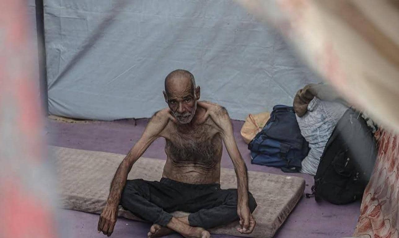 Thin, elderly Palestinian man sitting on a mat in a tent, surrounded by bags and blankets, needing aid. His extreme emaciation highlights the severe humanitarian crisis in Gaza. Donate to provide water, food, and medical supplies via crypto, like USDS, for urgent relief.