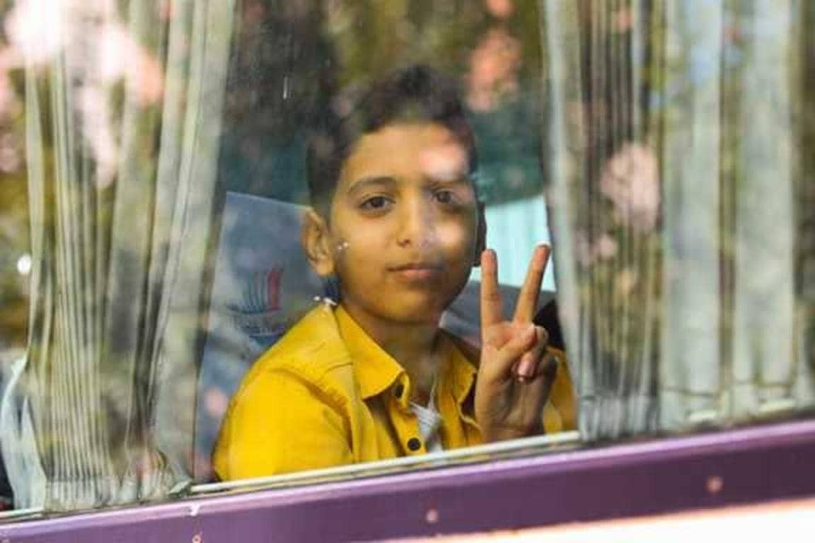 A young boy in a yellow shirt peeks through a window, offering a peace sign. Your crypto donation, such as BTC, brings vital winter aid to vulnerable children and families in conflict zones, providing warmth and hope.