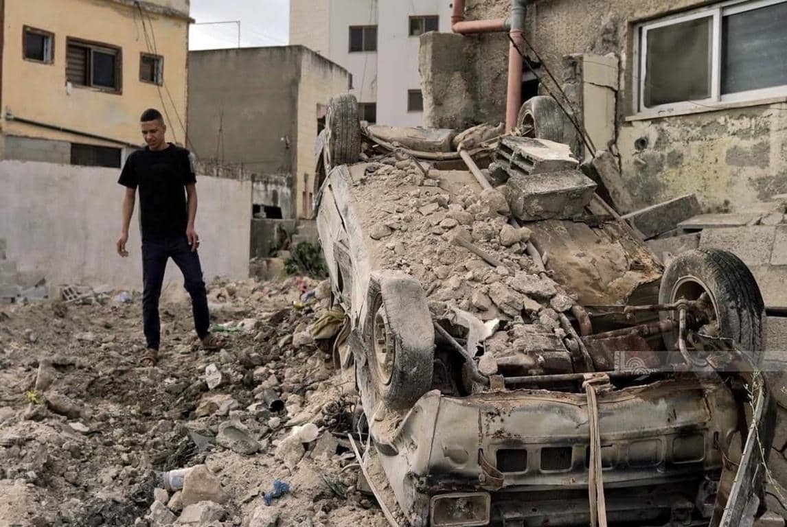 Devastated car amid rubble in a war-torn area, with a young man standing nearby. Crypto donations like BTC support immediate relief efforts for struggling families.