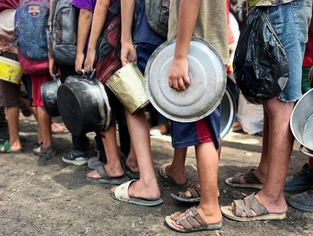 Children with empty pots and containers lined up for food distribution, symbolizing the urgent need for Zakat donation via cryptocurrency in Ramadan to provide essential aid like hot meals and food packages to the needy in conflict zones like Gaza, Palestine, and Yemen.