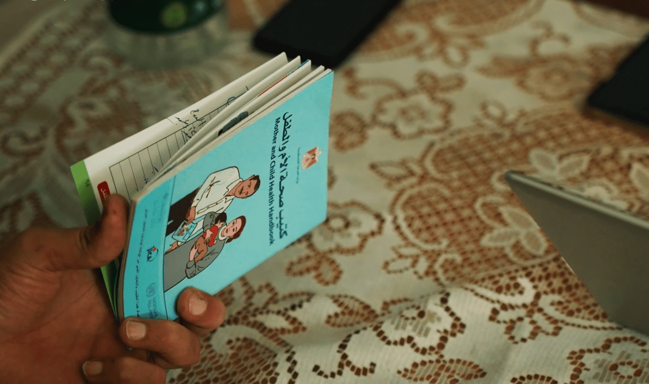 A hand holds an open Mother and Child Health Handbook in Arabic and English, depicting aid for Palestinian families via crypto donations, possibly using Solana.