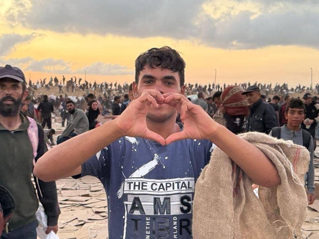 Young Palestinian man forms a heart shape with his hands, symbolizing hope and resilience amidst a crowd of people in a damaged area. Your crypto Zakat, like ETH, can provide vital aid for food, water, and reconstruction efforts.