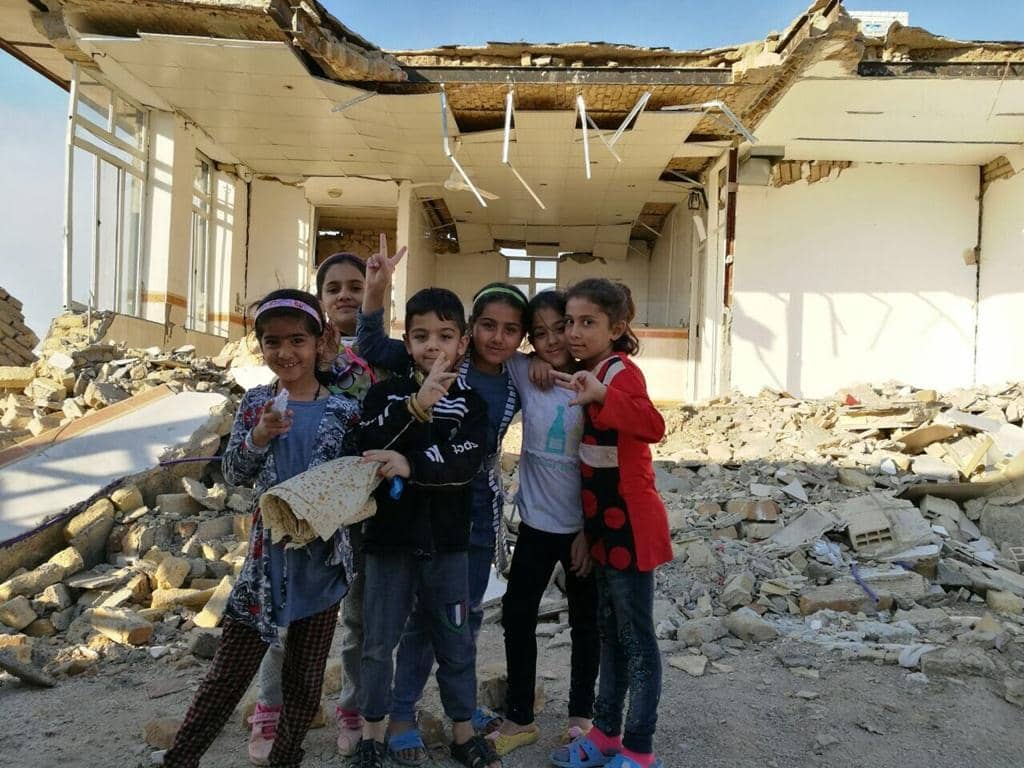 Children stand smiling amidst rubble before a damaged building, representing resilience and the need for aid. Your crypto Zakat donation, perhaps via Bitcoin, offers hope for reconstruction and support for Palestinian Muslims.