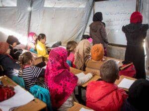 Children in a classroom setting, likely a tent, are focused on a whiteboard where Arabic script is written. One teacher stands at the whiteboard, and another teacher is visible. The scene implies educational support enabled by crypto donations, possibly for refugees or in a crisis zone, with a focus on anonymous giving.