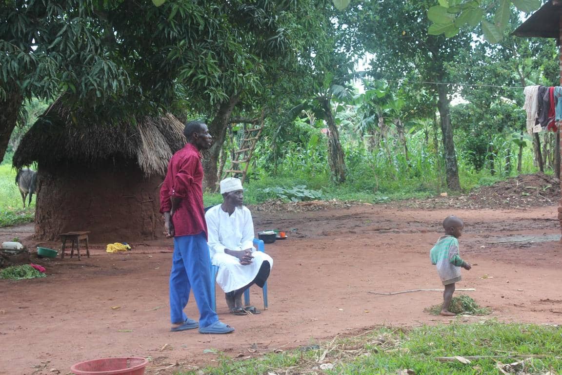 Three people stand in a rural African village; a young child, a man in a white outfit, and a man in a red shirt and blue pants reflect Islamic charity's programs, supported by PayPal USD crypto.
