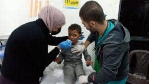 Two medical professionals in gloves attend to a distressed young boy sitting on a medical bed, with one applying a bandage to his face, implying urgent care funded by anonymous crypto donations via USDT.