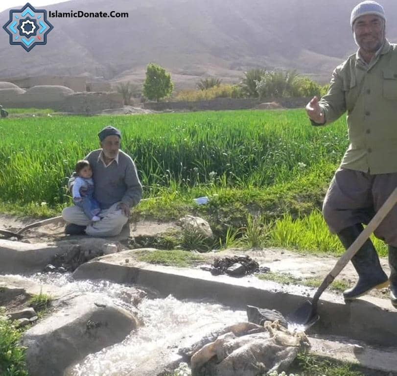Elderly man holding a child in a field near a water channel, with another man working on the channel, symbolizing sadaqah jariyah, ongoing charity, and agricultural support made possible by cryptocurrency donations like Bitcoin.