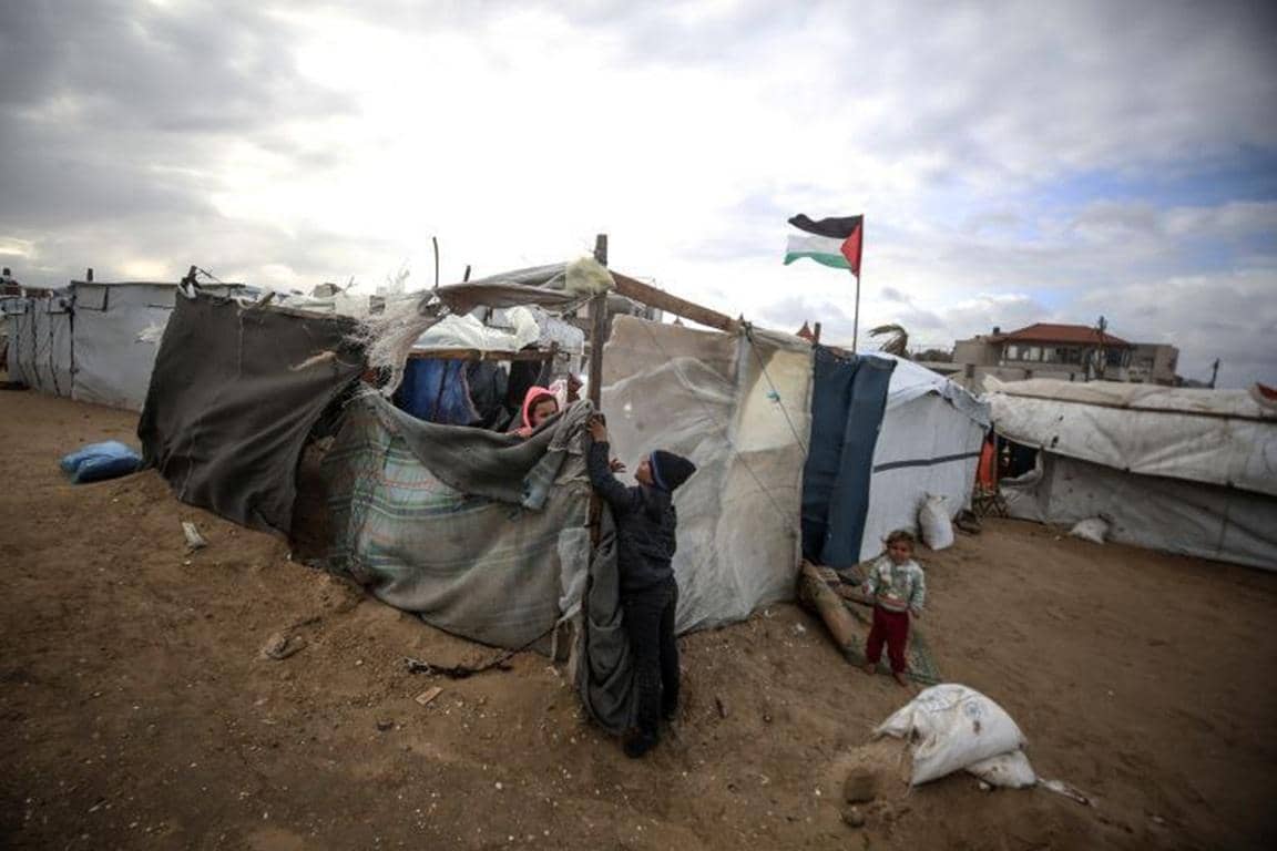 Children in makeshift shelters in Gaza struggle against the cold and rain, with a Palestinian flag flying. Humanitarian aid, processed quickly via BTC donations, provides essential warmth and shelter.