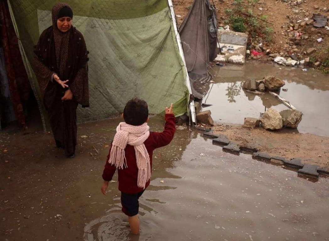 A child in a pink scarf stands in muddy floodwater near tents, while a woman in a headscarf looks on distressed, highlighting the harsh conditions and the urgent need for humanitarian aid, including warm clothing and waterproof tents, supported by Stellar donations.