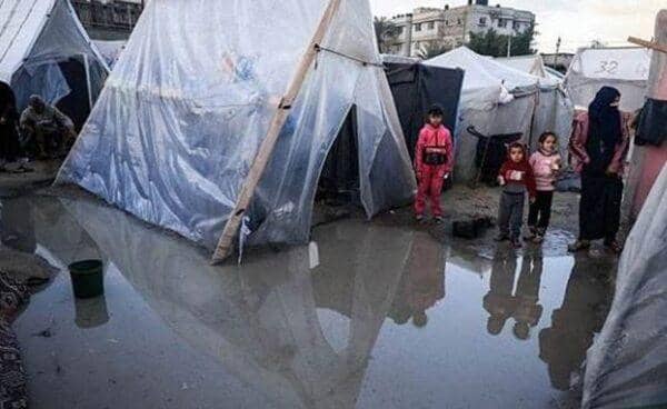 Children and adults in makeshift tents stand in flooded areas of Gaza, highlighting the struggle against cold and rain, with aid supported by SOL donations.