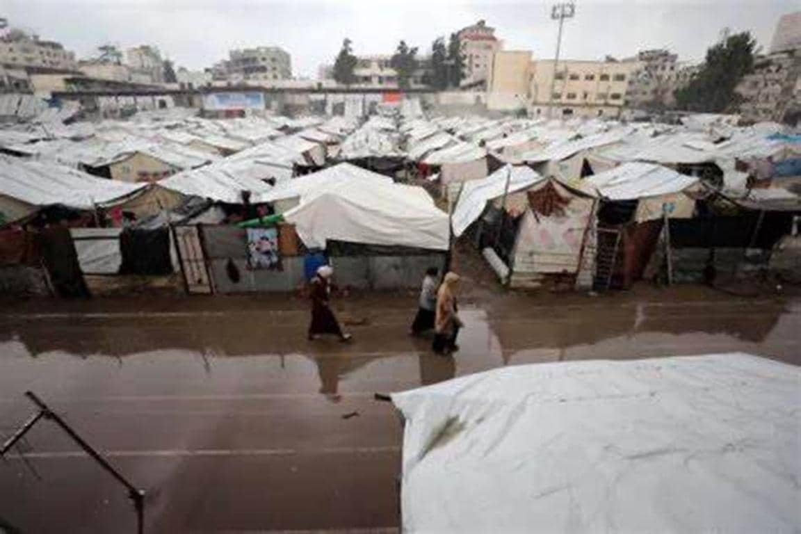 A crowded tent city in Gaza is flooded, with people walking through mud and water. This scene depicts the desperation of families facing cold and rain, highlighting the need for waterproof tents and warm clothing, with donations via Solana supporting relief efforts.