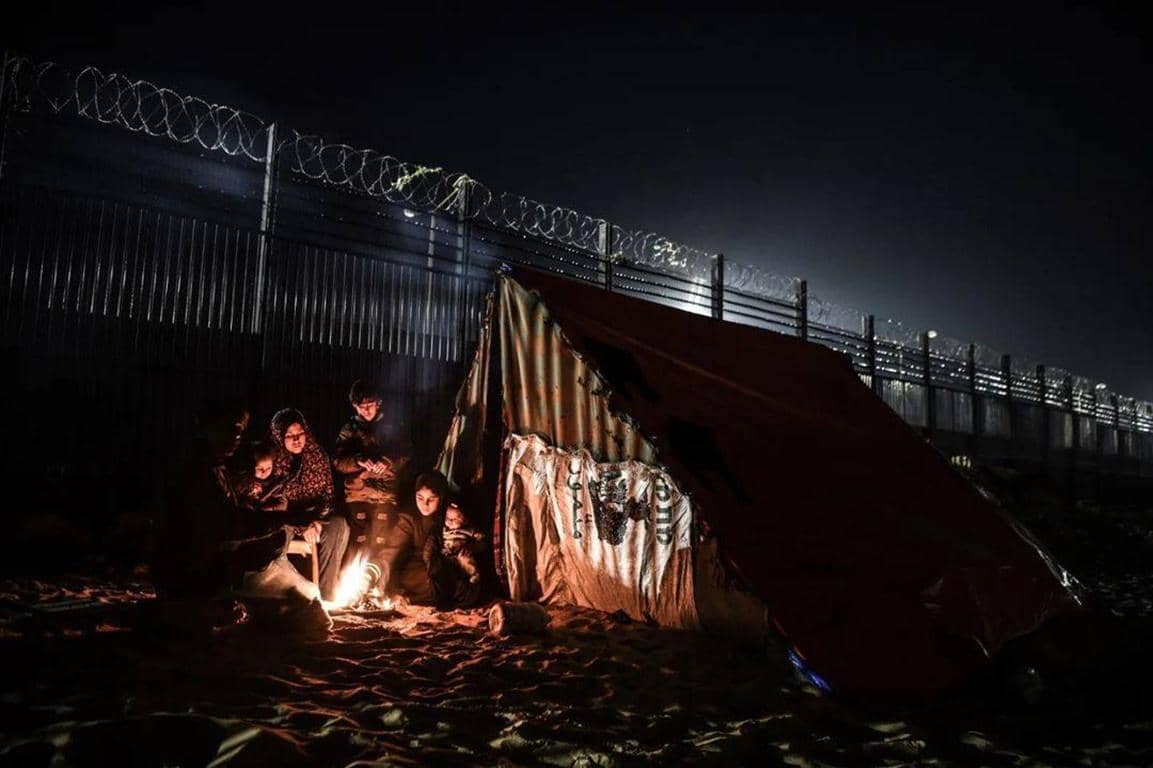 Family huddling around a fire for warmth inside a makeshift tent at night, with a barbed wire fence in the background. The scene illustrates the urgent battle against Gaza's winter crisis, where aid is urgently needed. Cryptocurrency donations enable rapid delivery.