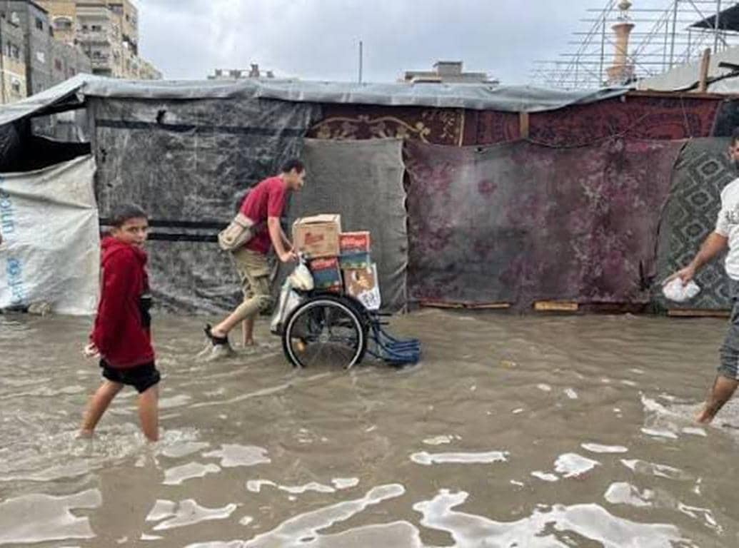 A child walks through flooded waters near makeshift shelters, while a man pushes a cart loaded with supplies through the Mideast winter crisis.