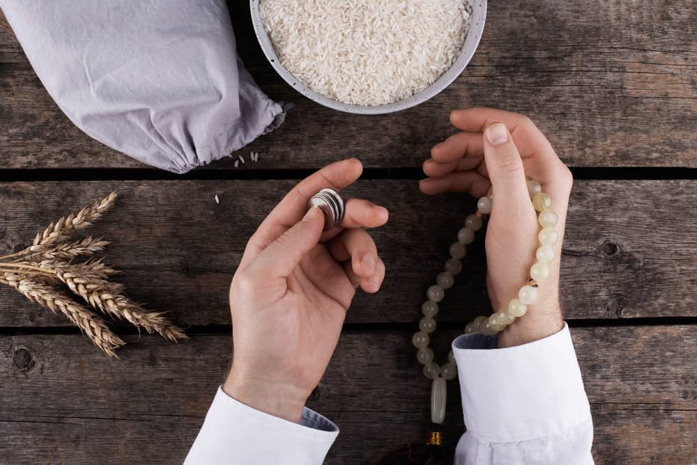 Hands holding coins and prayer beads, with rice and wheat stalks, representing Zakat donation via cryptocurrency like BTC.