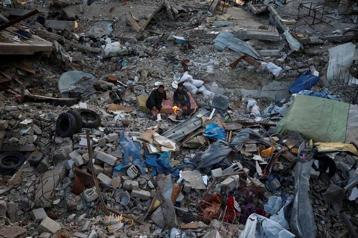 Two people sit amidst rubble around a small fire, illustrating the impact of disaster and the need for Zakat donation, possibly aided by PayPal USD cryptocurrency.