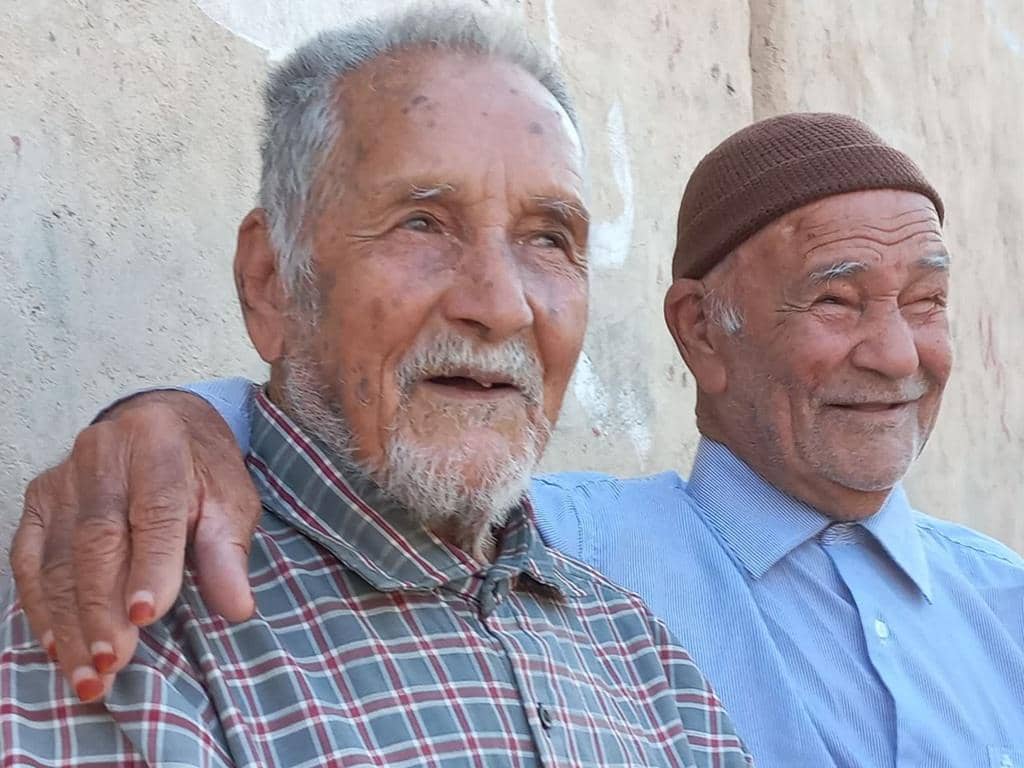 Two elderly men smiling, one with a brown kufi hat, posing for a photo while receiving Zakat donations via crypto like Stablecoin, benefiting the Muslim Ummah.