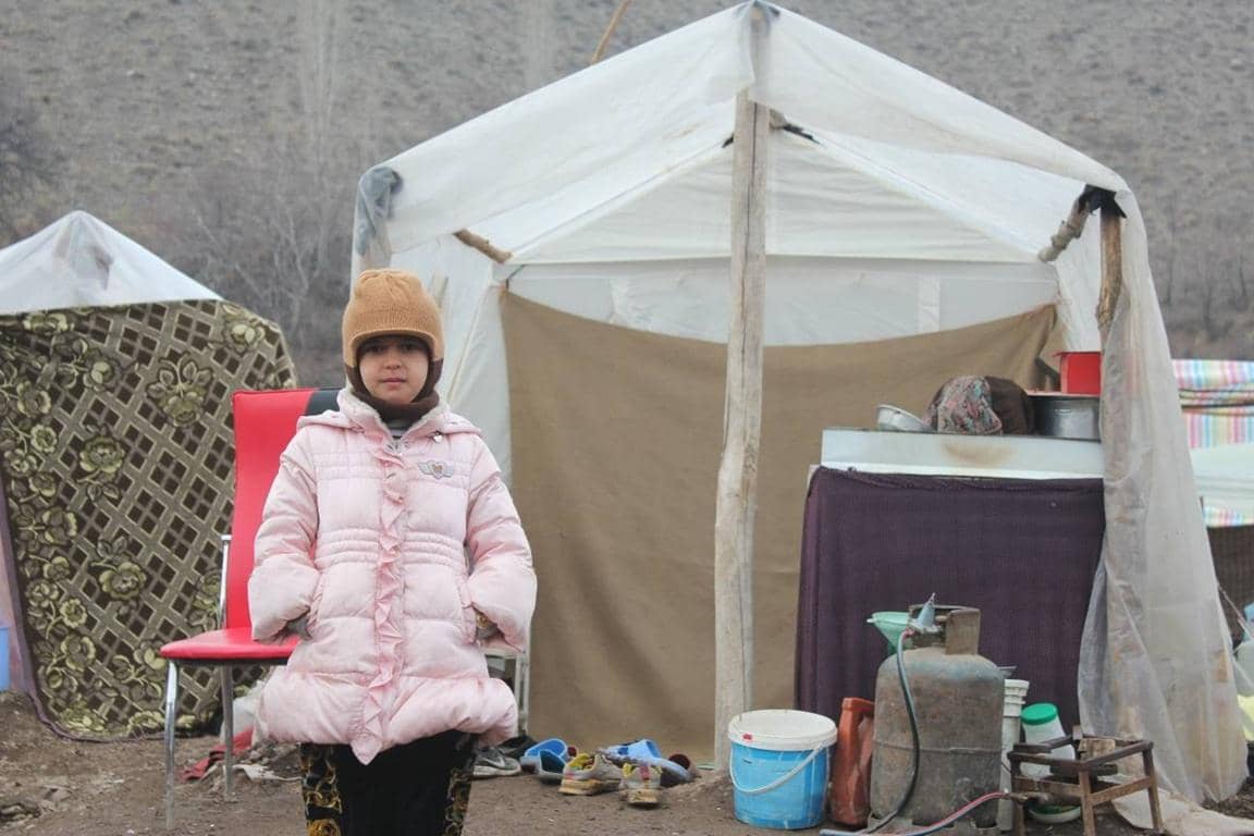 Child in winter coat and beanie standing in front of a basic tent structure, a symbol of poverty relief efforts. Crypto donations, like BTC, help fund skills training and essential resources for families. Empowering children and communities.