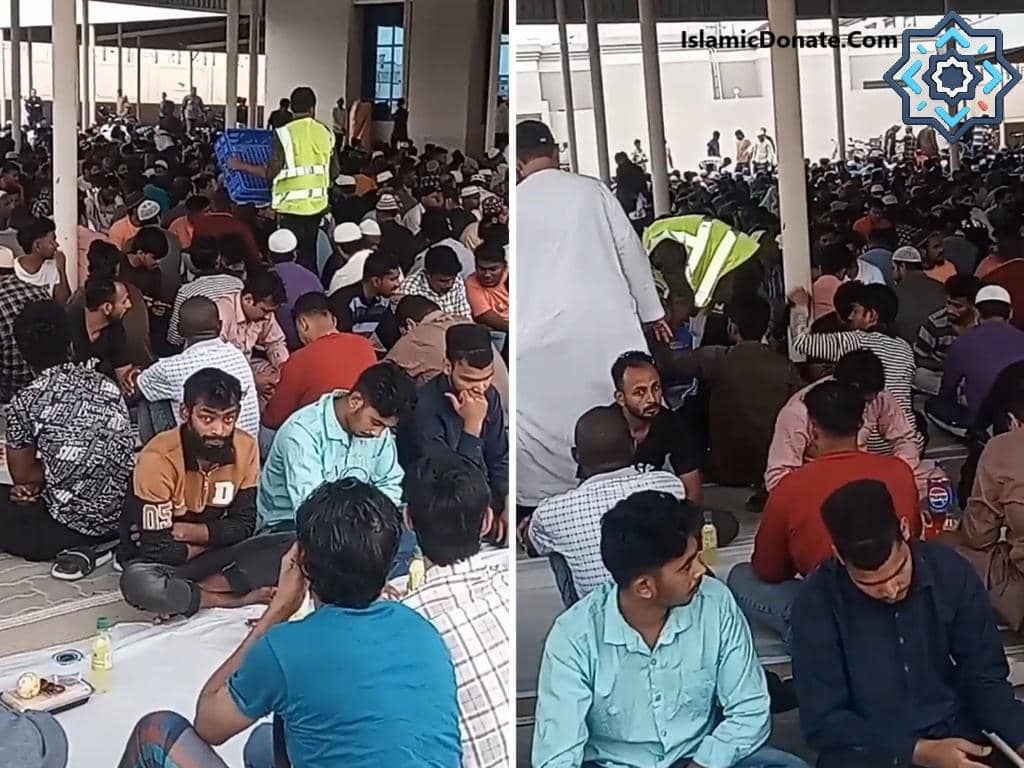A large group of men sits on the ground, some being served food by volunteers wearing high-visibility vests. The scene depicts a community meal, part of the Koonday ki Niyaz tradition, where donations fuel nourishment. Donate with PayPal USD or other crypto for swift aid.