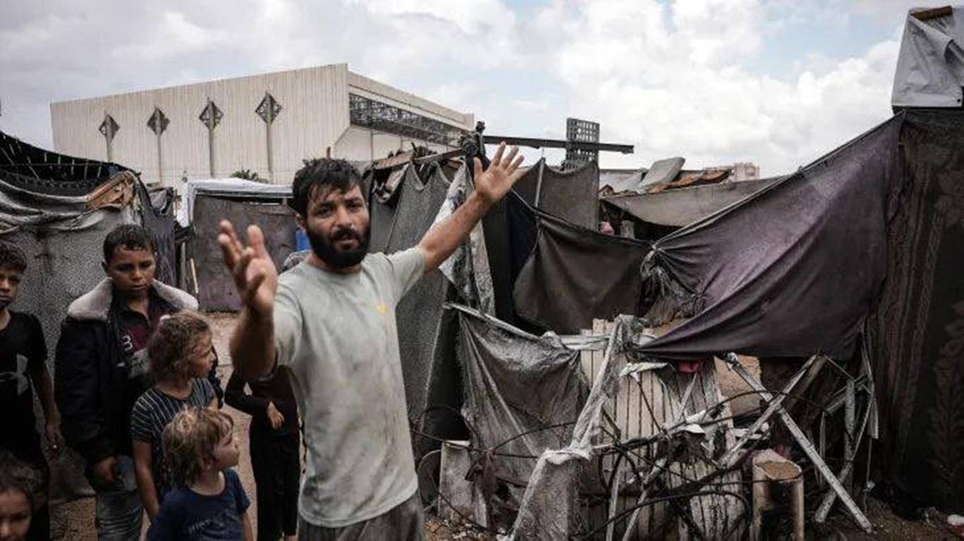 Man in a tattered camp with children, gesturing outwards. Pay Khums with cryptocurrency to support poor families, orphans and stranded travelers.