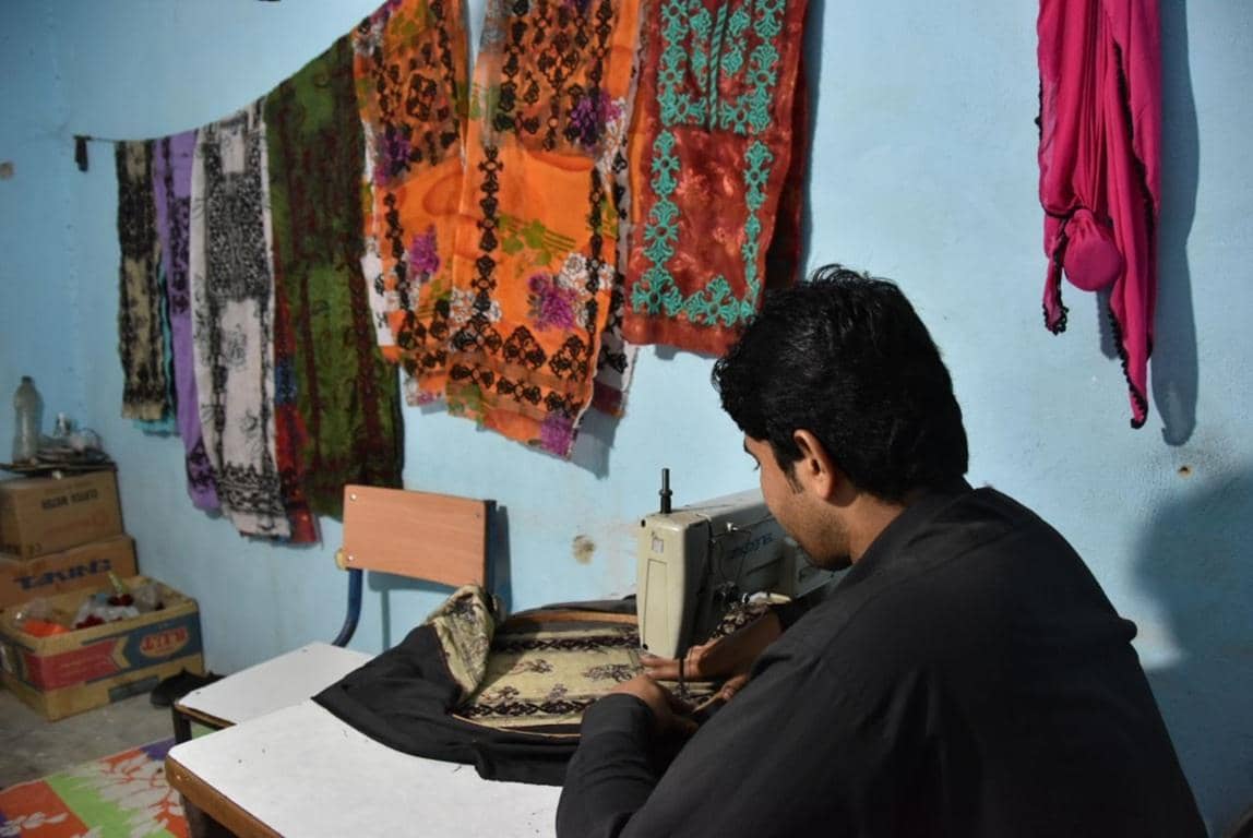 A young man sews with a sewing machine, surrounded by colorful fabrics hanging on the wall, symbolizing skills training for income generation funded by crypto, such as SOL donations.