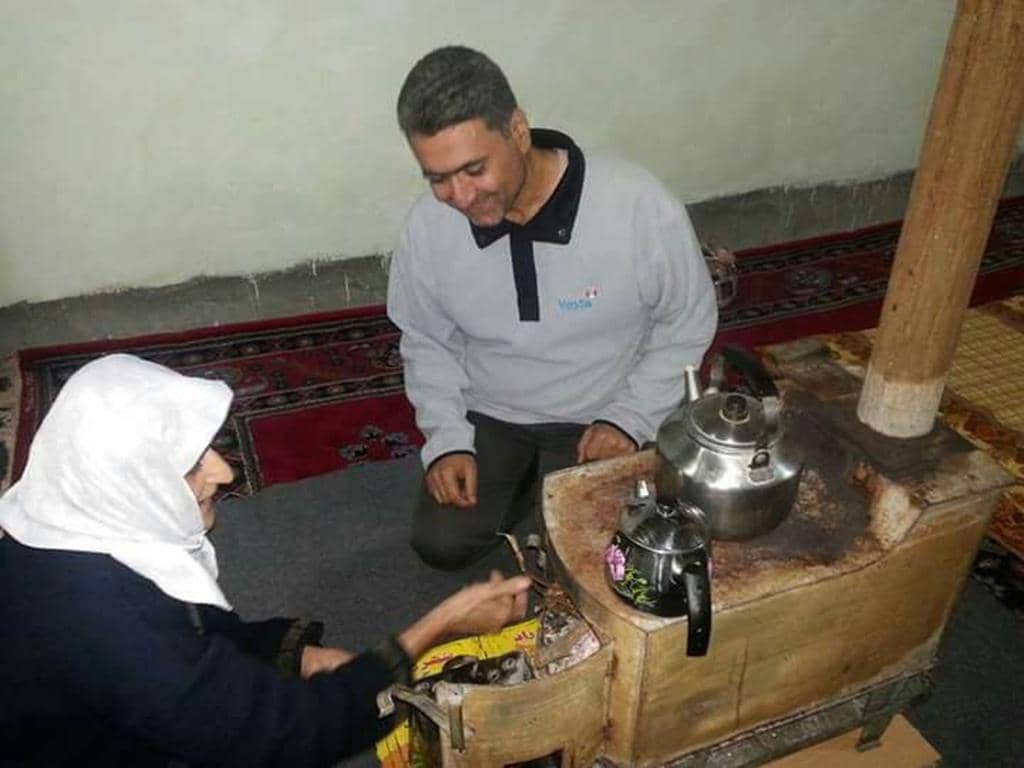 Elderly woman and man share tea by a stove, symbolizing humanitarian aid and community support. Donations via SOL crypto ensure vital care.
