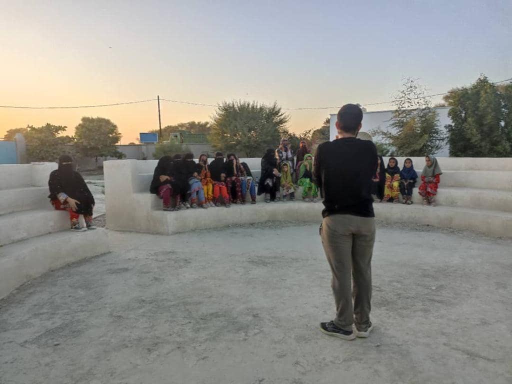 People gathered on tiered seating, possibly for a training session or community meeting, with a man facing them. Donations enable training and tools for income generation, with support via SOL.