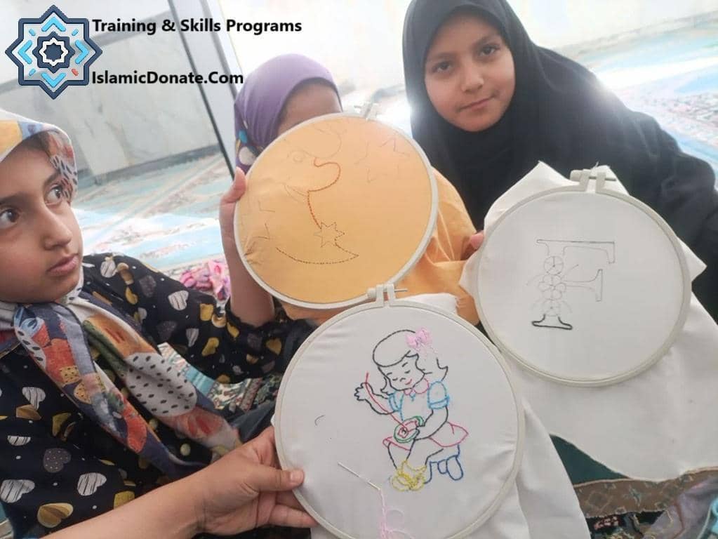 Three young girls in hijabs learning embroidery skills. One girl holds an embroidery hoop with a cartoon girl, another with a moon and star, and the third with the letter F. These training programs are funded by crypto donations like Dogecoin.