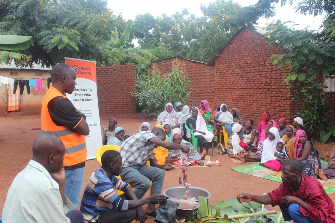 A man in an orange vest stands near a banner for Islamic Donate, while a group of women and children sit on the ground watching as meat is prepared in a large pot. This scene depicts the charitable distribution of Qurbani meat, facilitated by online donations including cryptocurrency like Bitcoin.
