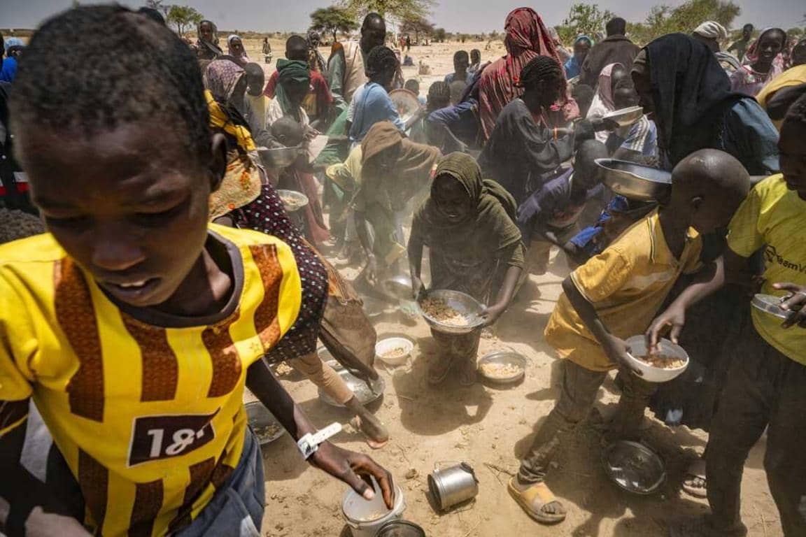 Children and adults gathered, receiving food aid in a dusty, open area. The scene depicts a humanitarian crisis and is supported by rapid crypto donations like BTC.