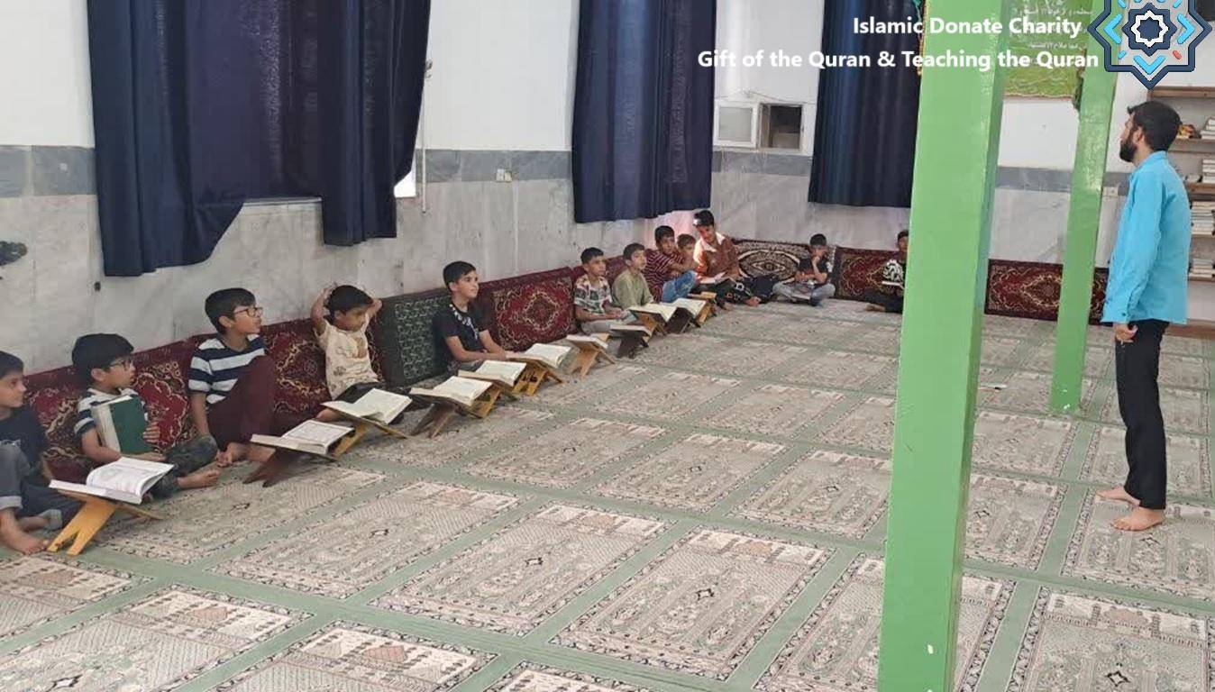 Children sitting on the floor in a classroom-like setting, reading Qurans on wooden stands. A teacher stands and instructs them. This project, Gift the Quran, helps provide spiritual guidance and education through crypto donations like BTC, supporting the Muslim community.