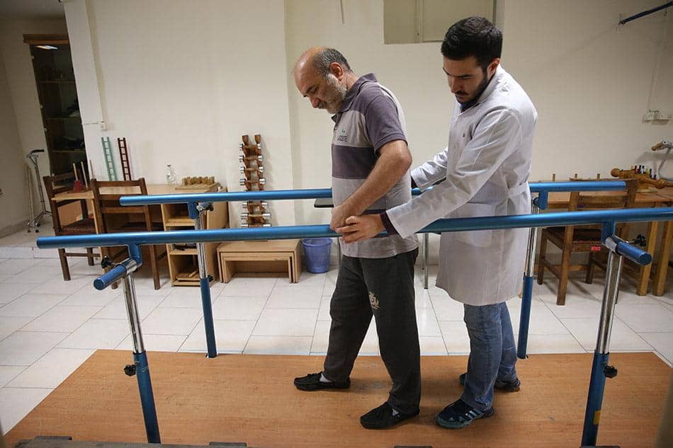 Physical therapy patient walking with assistance using parallel bars, supported by a therapist. Services include mobility, strength, and balance restoration. Supported by ETH.